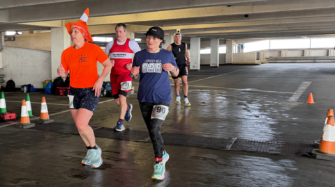 Runners in motion in a car park. They are dressed in running gear, with one runner wearing a fabric orange traffic cone on his head. 