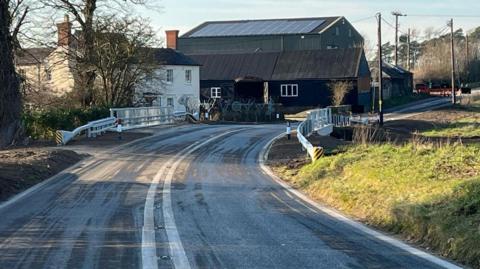 A view of the Bridge Farm Bridge in Stowlangtoft. The road is open. Buildings can be seen on the other side on the left hand side of the road. 