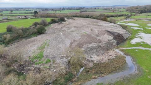 A picture of what appears to be a large illegal waste tip in the countryside. The site is flanked by green field on one side and flood plain on the other. The aerial view seems to show rubble and waste in the field. 