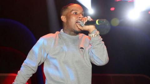 Tory Lanez performs on stage. He is wearing a grey jumper and is singing into a mic. He stands in front of black background. 