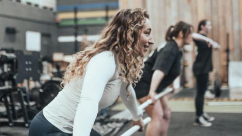 A woman in athletic clothing practices a lifting movement with a white training bar inside a gym. She is positioned in a partial squat with the bar held horizontally in front. Next to her, to the right, are two other women who are also holding training bars and performing similar movements. The gym space includes weight racks, exercise machines, rubber flooring, and high ceilings with ropes hanging down.