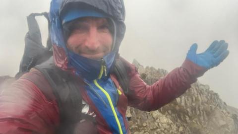 Fell top assessor Paddy Cave at top of Helvellyn in heavy rain and poor visibility. He is showing the strength of the high winds at the summit. He is wearing a maroon and blue waterproof which is soaked through.