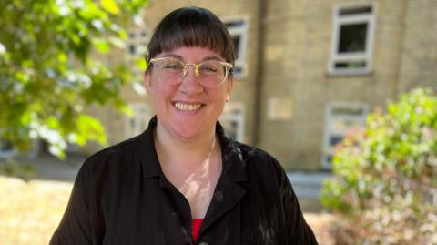 She is wearing a red top paired with an oversized black cardigan. Her dark brown hair is in a fringe and she is is wearing clear framed glasses, highlighted in gold.
 She has one hand on her hip.