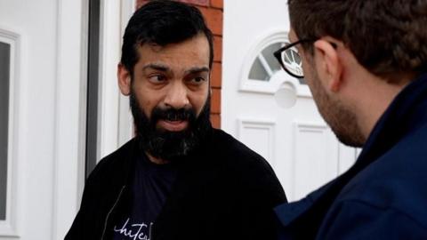 Mr Prabhakar, who has a beard and is wearing a black jacket, stands outside a while front door. With his back to the camera, a BBC journalist questions Mr Prabhakar. 