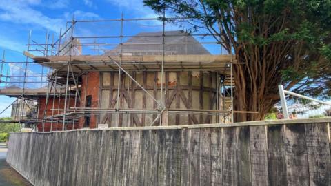 Bramcote Tennis Pavilion in Scarborough surrounded by scaffolding. The 19th Century pavilion is a red-brick building with Tudor style wooden cladding around one section. A wooden slatted fence stands in front of it, and a large Conifer tree is to the left.