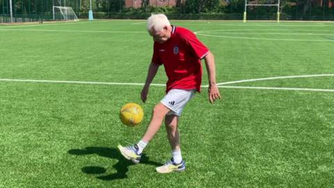 A person is standing on a green sports field, keeping a yellow football in the air with one foot.
The field has goalposts, markings, and is surrounded by trees and red-brick buildings in the background.
The person is wearing a red England shirt, white shorts, and trainers on a bright sunny day.