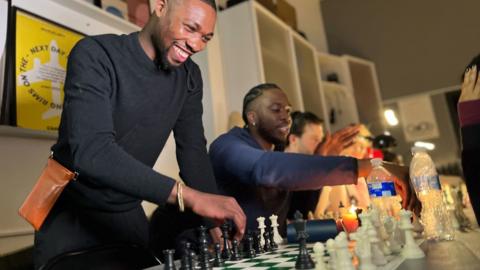 Kojo Dwimoh smiles as he plays chess. He is surrounded by others playing the game. 