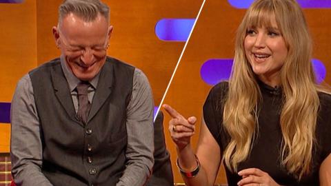 Bruce Springsteen sits on Graham Norton's orange sofa and looks down laughing. He is wearing a light grey shirt and dark grey waistcoat. He is alongside Jennifer Lawrence, also sat on the sofa, who is pointing with one finger and is wearing a black dress.