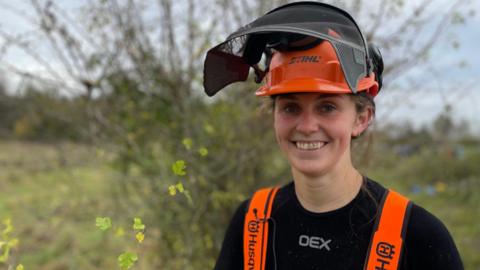 A woman wearing an orange hard hat and visor, with orange braces, smiles at the camera in front of hedges.