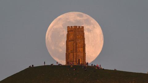A huge pink hued Moon sits in a grey sky, behind the tower on Glastonbury Tor with people milling around at the base