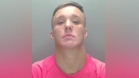 Mugshot of Brandon Welch, he has short fair hair and is wearing a pink t shirt.