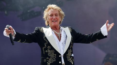 Rod Stewart is standing on stage wearing a black jacket with stitched flowers. He is spreading his arms towards the crowd and has a microphone in hand. His shirt has a low neckline and he is wearing about three necklaces.