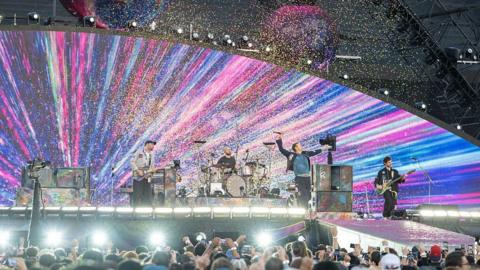 A live concert with four band members performing on stage under bright multicoloured lights and falling confetti.