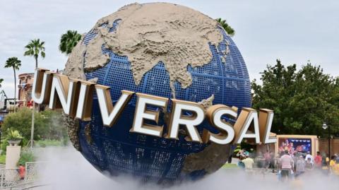 A large model of a globe stands within a theme park. Around the Equator is the logo "UNIVERSAL" in capital letters. There are some people in shot, along with some palm trees.