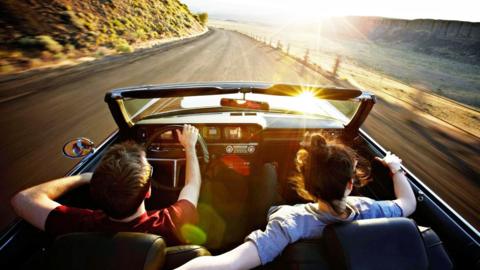 A young man and woman in a convertible car driving along a mountain road towards the sunset. She is driving and has her arm around the back of his seat.