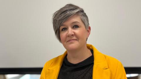 Deputy Haley Camp - A lady with short black and grey hair. She is wearing a yellow jacket and a black t-shirt. 