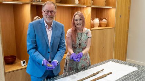 Paul, who wears a light blue suit, stands with Emma who has pink hair and wears a light blue and pink dress. They are both holding artefacts and wearing purple gloves. The swords are on a table in front of them. 