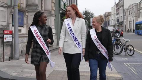 (L-R) Krystel Vida, Alice Cutler and Charlie Cobb, all wearing sashes and smiling are talking and walking towards the camera on a pavement in a built up city
