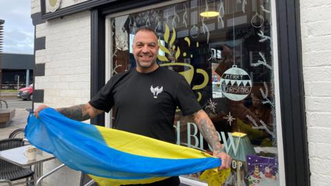 A man with short dark hair and tattoos on his arms stands in front of a shop window. The shop is decorated for Christmas and the man is holding a Ukrainian flag.