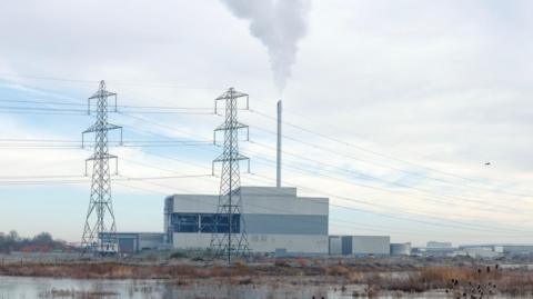 A white building with a single chimney that releases a white cloud into the sky. The foreground is brown marshland and two pylons can be seen in front of the plant. The sky is partially cloudy. 