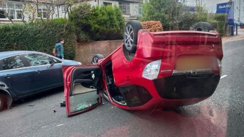 A red car that has been flipped over and is balancing on its roof. In the background are houses. 