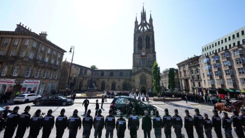A funeral cortege arrives at Newcastle Cathedral as rows of police officers stand in formation, with mourners gathered outside the church during a ceremonial service. It is a sunny day. Attendees stand on the edge of the square in front of the cathedral, with clergy waiting for the cortege in the centre of the plaza.