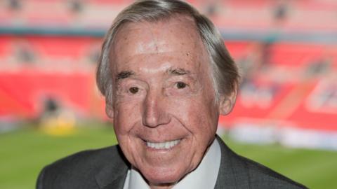 A head and shoulders shot of a smiling elderly man with fair hair combed over and long around his ears. He has brown eyes. The red seats and pitch of Wembley Stadium can be seen blurred in the background.