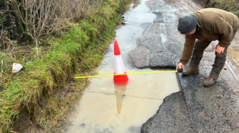 A man holding a measuring tape is bending over a pothole full of rain in Bridewell Lane, Oxfordshire. There is a red and white cone in the water. The single-track road has a grass band on one side. The man is wearing a green jumper and trousers with a grey hat and brown boots.