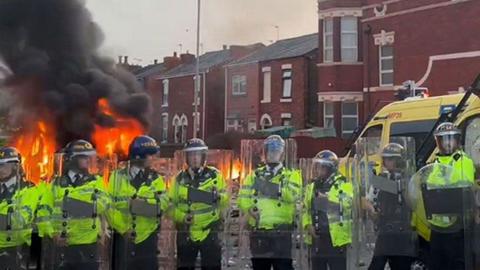 Line of riot police in high vis clothing holding riot shields outside mosque where one police van is on fire