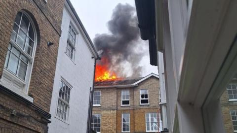 Flames and heavy smoke coming from the roof of a brick building in a built-up area.