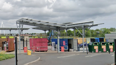 The entrance to Pallion Household Waste and Recycling Centre in Sunderland. There are a number of red, blue and green shipping containers in different sizes, surrounded by parking spaces. A row of green trees is visible behind the facility, with the Northern Spire bridge further back.