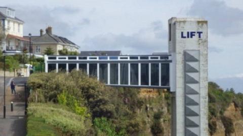 Grey column running down cliff with glass walkway connecting to a path and road with buildings alongside.
