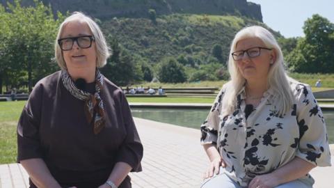 Two women sit by a plaza with a water feature and hill in the background. Cynthia Guthrie wears a brown top and dark glasses, and Allison Graham wears a patterned white and black shirt with dark glasses. Both have fair hair 
