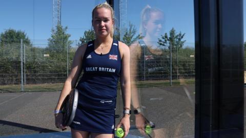 A woman in a blue tennis top and skirt, standing in a glass padel court. She is holding a ball and her padel racquet. She has blonde hair which is tied up in a ponytail and is smiling. 