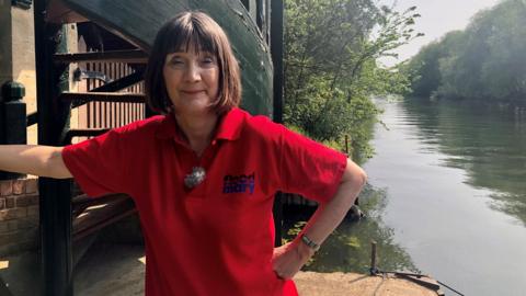 A woman with short brown hair and wearing a red polo shirt. She's standing in front of a river with a hand on her hip and it's a bright sunny day.