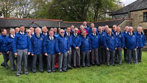 A group of around 25 men from the Minehead Male Voice Choir, all wearing shirt and tie, with blue coats and grey trousers. They're gathered on grass in front of a brick building all looking towards the camera.