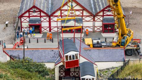 Two red and white carriages with grey curved roofs are hoisted into position on vertical railway track by a yellow crane. Workers in orange hi-vis clothing and bystanders watch in front of a red and white building with the beach behind.