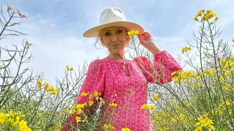 A woman wearing a pink dress and a hat. She is pictured in a field with yellow flowers around her.