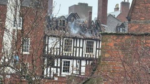 A black and white building with white smoke coming out of its roof. the roof has been on fire and is charred with the beams exposed