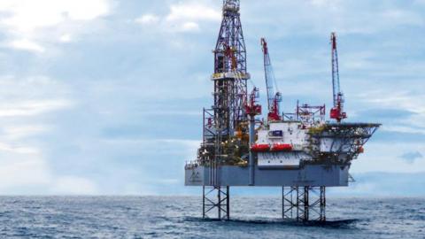 A rig sits in the sea protruding above the horizon with a blue sky with clouds. Three red cranes and a tall skeletal metal structure rise high from a sturdy grey deck.