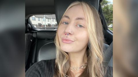 Kelly has taken a selfie in a car with black leather seats. Her seatbelt is covered by long, wavy brunette hair which has blonde dye throughout. She is wearing a black jumper with a striped pattern. A silver necklace made of a string of heart shapes rests on her neck. Kelly, who has green eyes, is pouting and has multiple piercings, including a silver stud in the centre of her upper lip. She has two silver hoops on her outer nostrils and a septum piercing, which has two pyramid shaped ends, one coloured pink and the other blue.  