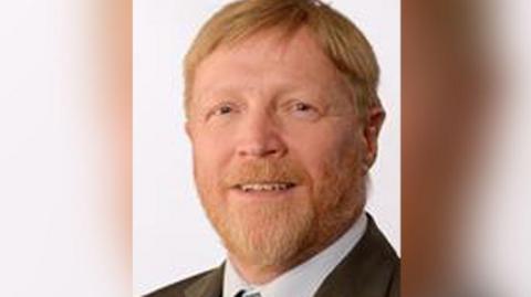 Councillor Stephen Thompson with red hair and beard wearing a brown suit with white shirt and tie. He is smiling.
