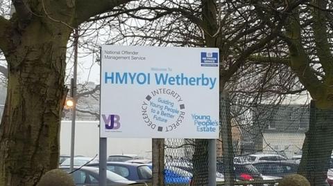 A sign for HMYOI Wetherby stands in front of trees and a wired fence.