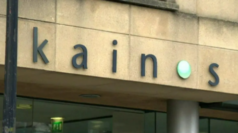 A shot of the exterior of Kainos's building in Belfast. The logo 'Kainos' written in lower case is in black with a green circle dot replacing the O.