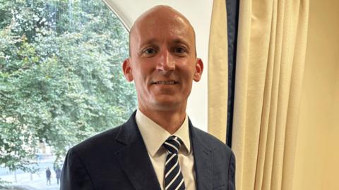 Sean Gaul is wearing a blue suit, white shirt, and blue and white striped tie. 