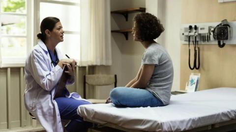 A woman sat on a bed in a doctor's surgery talking to a doctor sat opposite her in a chair