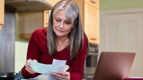 Woman looks over bills with a neutral expression. She stands in front of a laptop.