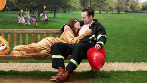 A man in a fireman’s uniform sits on a park bench while a woman lies on the bench next to him and lovingly looks up at him.