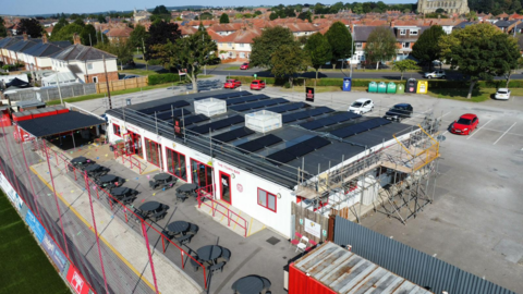 A white clubhouse building with red-framed windows and a roof covered in solar panels at Bridlington Town FC's stadium. Part of the pitch can be seen to the left, while a car park can be seen in the background.