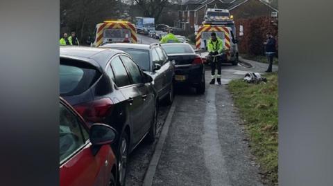 Four cars parked on a road with a tow-away truck loaded with cars ahead of them. A man in high-vis jacket is making notes. Three other men in high-vis jackets can be seen nearby with another tow truck.